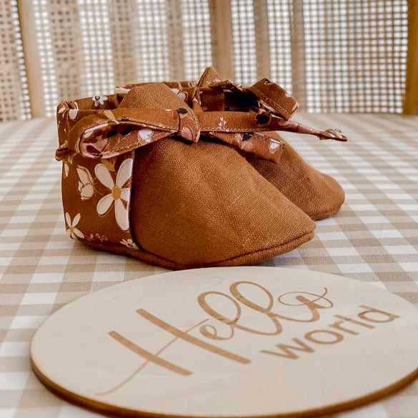 a Pair of Brown Linen baby booties with floral backing on a baby bassinet with an announcement disc