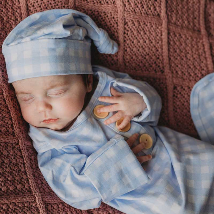 Baby boy laying on a newborn knitted blanket by snuggly jacks wearing a newborn knotted gown. Snuggly Jacks Canada cornflower Gingham Knot Gown: Cotton Jersey Stretch Fabric, check blue fabric and matching beanie