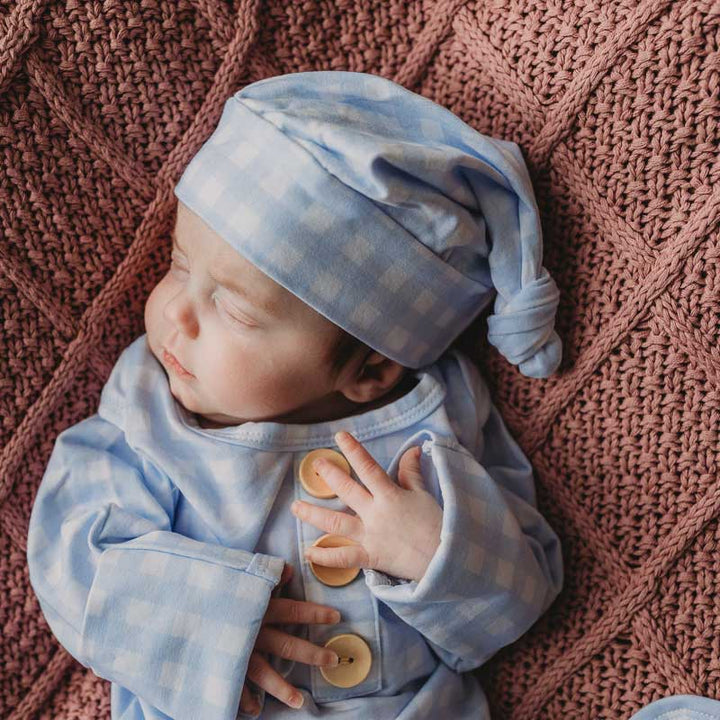 Baby boy laying on a newborn knitted blanket by snuggly jacks wearing a newborn knotted gown. Snuggly Jacks Canada cornflower Gingham Knot Gown: Cotton Jersey Stretch Fabric, check blue fabric and matching beanie