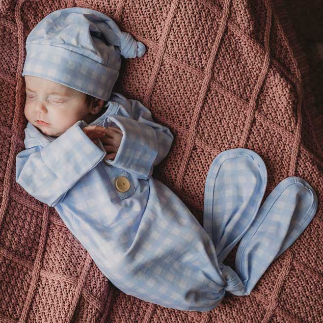 Baby boy laying on a newborn knitted blanket by snuggly jacks wearing a newborn knotted gown. Snuggly Jacks Canada cornflower Gingham Knot Gown: Cotton Jersey Stretch Fabric, check blue fabric and matching beanie