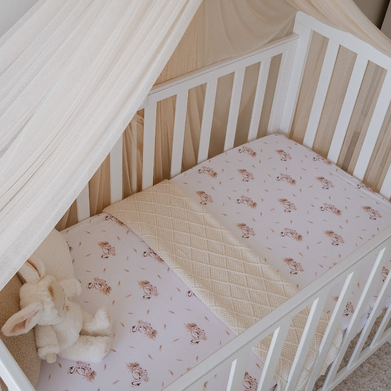 White crib with a tule canopy, plush bunny, knitted cotton blanket layered across a crib sheet with swans on it.