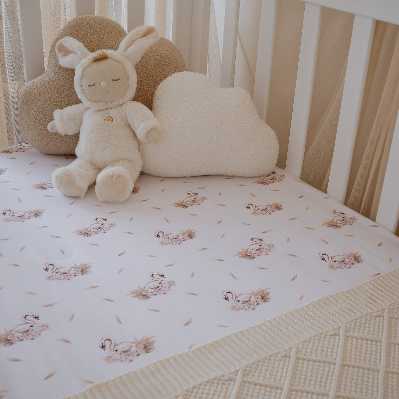 Adorable plush toy sitting in the conner of a crib set with a swan printed fitted crib sheet from snuggly jacks Canada.