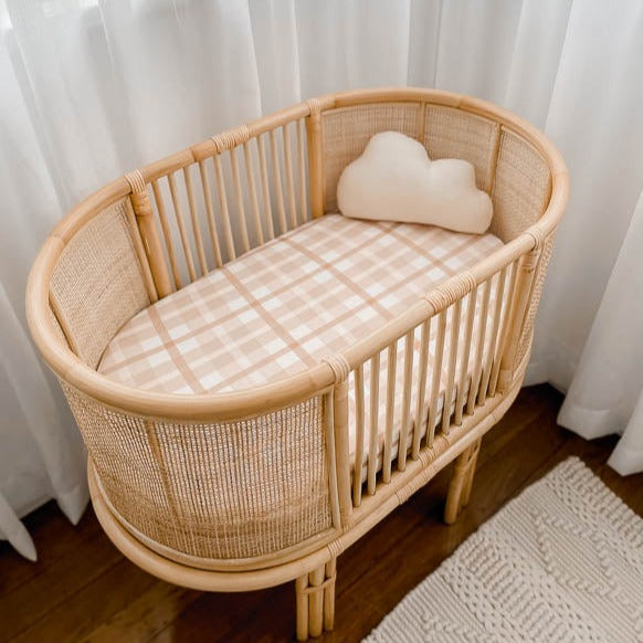Rattan bassinet in the corner of a room with soft defused light helping show off the soft browns of a cotton bassinet sheet and a cloud pillow.
