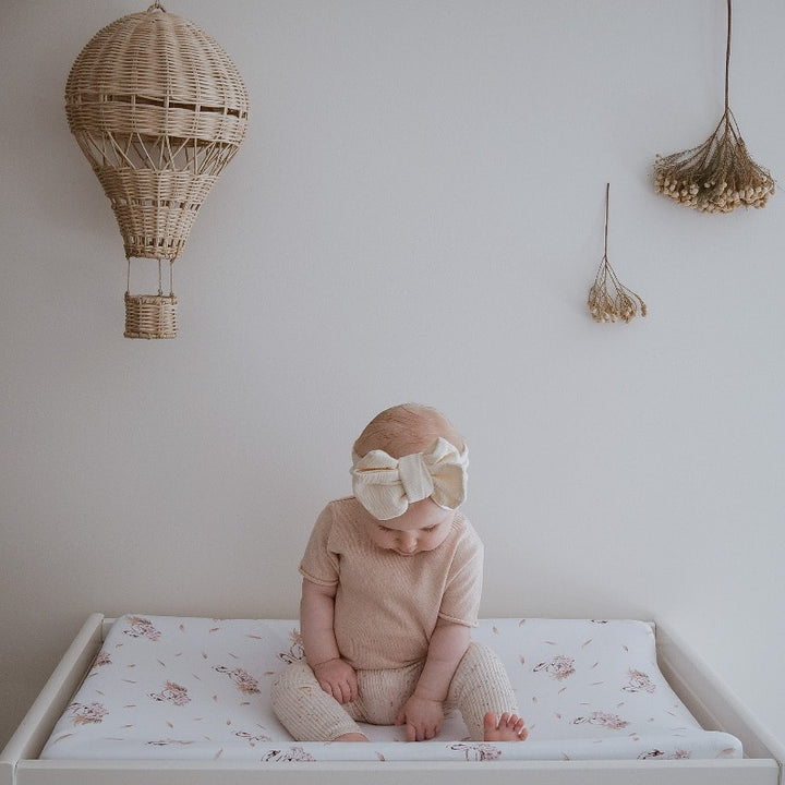 Cute baby sitting up on a change table wearing a linen bow, there is a rattan hot air balloon hanging on the wall beside her.