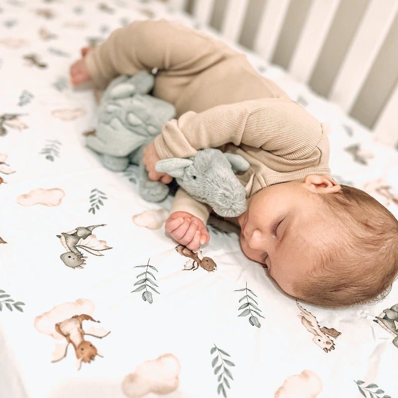 The cutest of babies sleeping while snuggling a plush toy in a crib. The sheet is white with little dragons, green leaves and soft brown clouds all over it. 