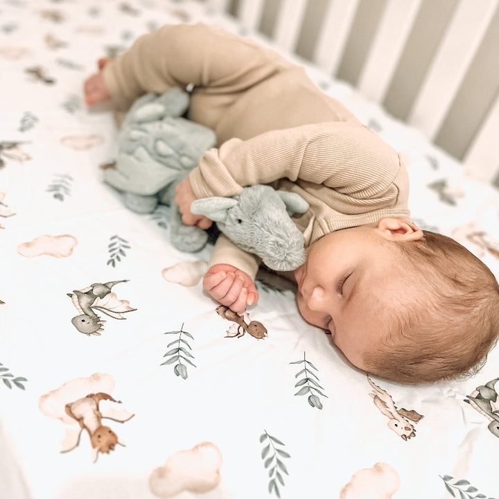 The cutest of babies sleeping while snuggling a plush toy in a cot. The sheet is white with little dragons, green leaves and soft brown clouds all over it. 