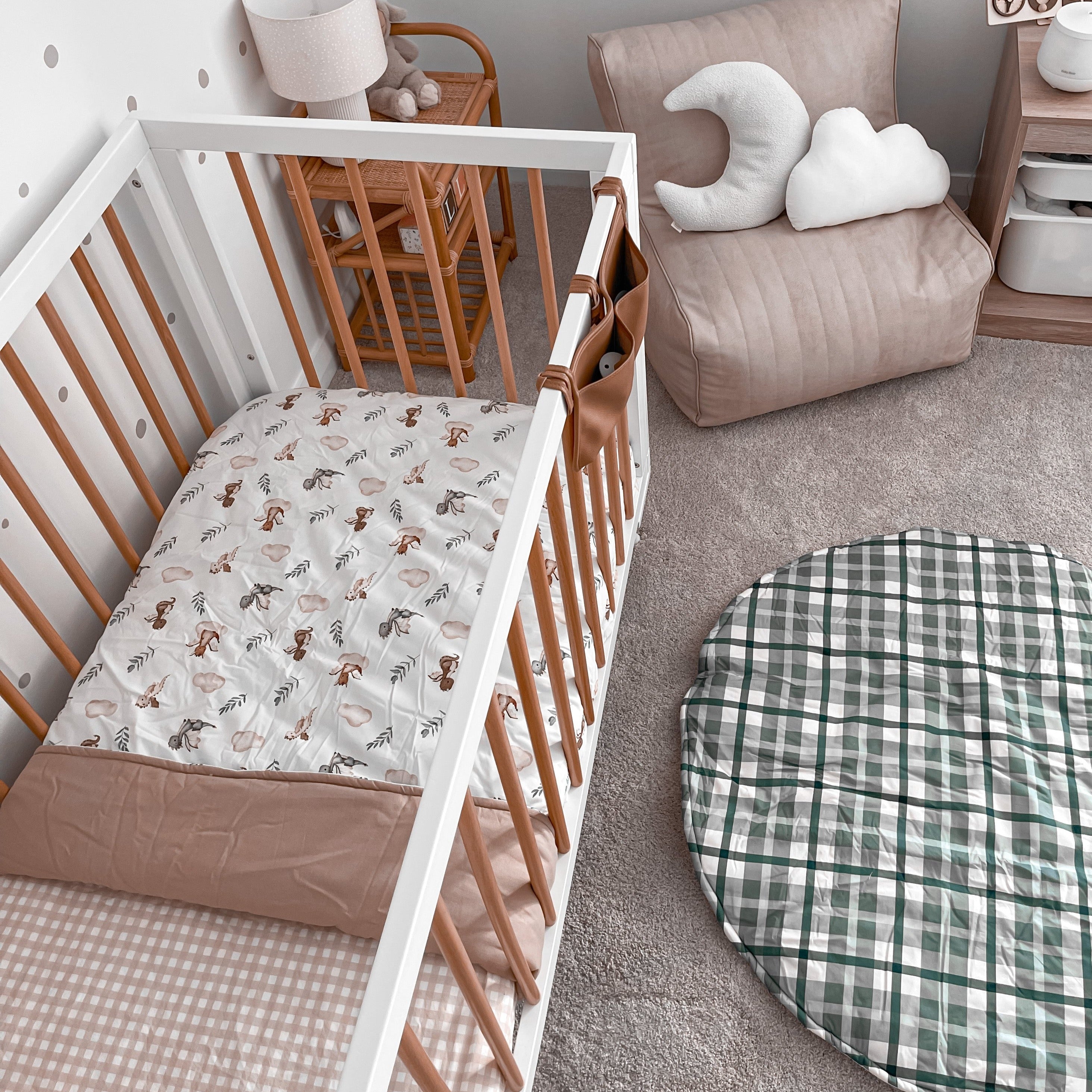 Modern nursery set with a blue plaid cotton playmat, soft chair in one corner, a rattan shelf and a pine crib in the bottom corner made up with the a cotton fitted sheet and a cotton crib quilt.