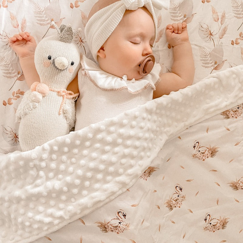 Adorable baby sleeping on a pattern with pastel colored leaves with a dimple dot minky blanket laying over her and her plush toy.