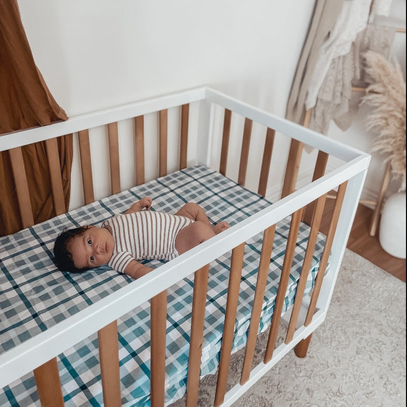 A Pine crib with white accents housing a baby looking at the camera while relaxing on a plaid patterned cotton crib sheet