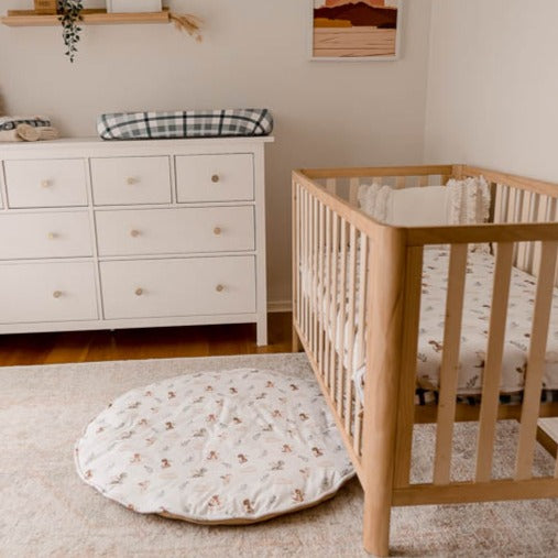 Wide shot of a nursery with soft colours featuring a pine crib, cotton play mat, change pad cover and a cotton fitted sheet all with the mystique dragon print on it.