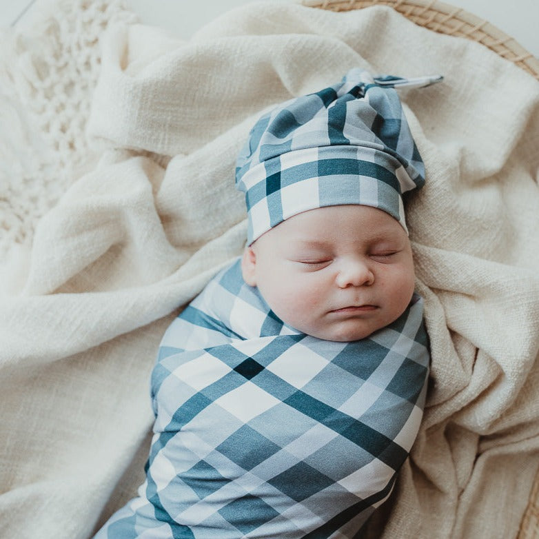 Sleeping baby laying in a moses basket all wrapped up in a snuggly stretch wrap and wearing a matching blue plaid beanie.