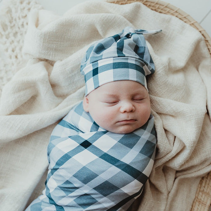 Sleeping baby laying in a moses basket all wrapped up in a snuggly stretch wrap and wearing a matching blue plaid beanie.