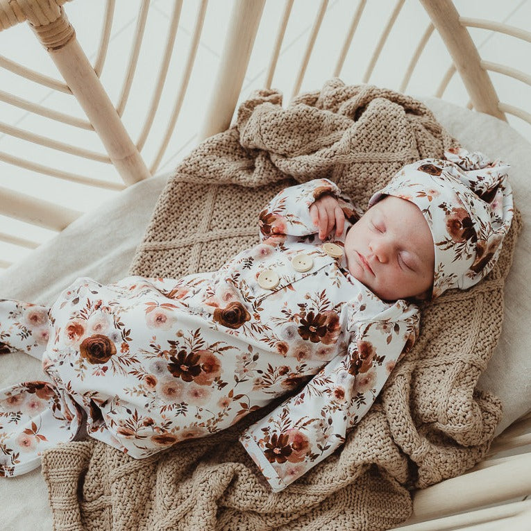 A sweet little baby laying on a cotton knitted blanket and wearing a maroon floral beanie and knotted gown
